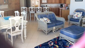 A spacious kitchen with wooden cabinets, a dining area with white chairs, and wicker furniture with blue cushions.