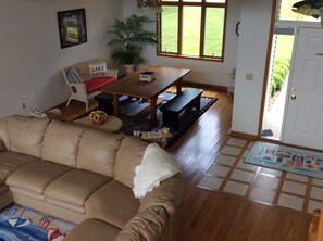 A living room with a beige sofa, a wooden dining table with benches, a potted plant, and a door leading outside. - Create your family memories at our nautical lakeside home! (Ashville)