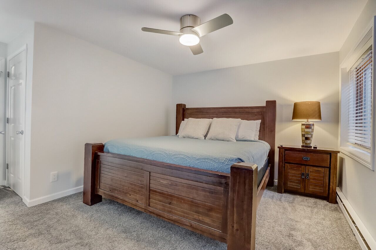 Un dormitorio con una cama de madera, un ventilador de techo, una mesita de noche con lámpara y una ventana con persianas.