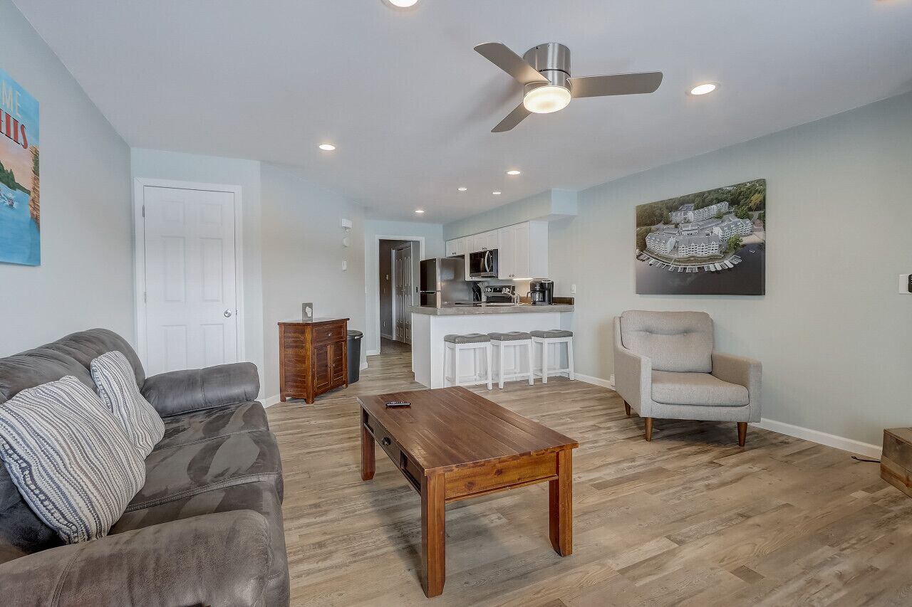 A living room with a sofa, a coffee table, a ceiling fan, and a kitchen area with a bar and stools.