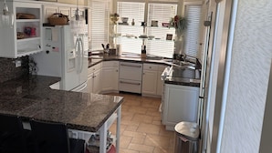 A kitchen with white cabinets, granite countertops, and a central island. There are windows with blinds, a ceiling vent, and a trash can. - Beautiful ocean retreat (Morro Bay)