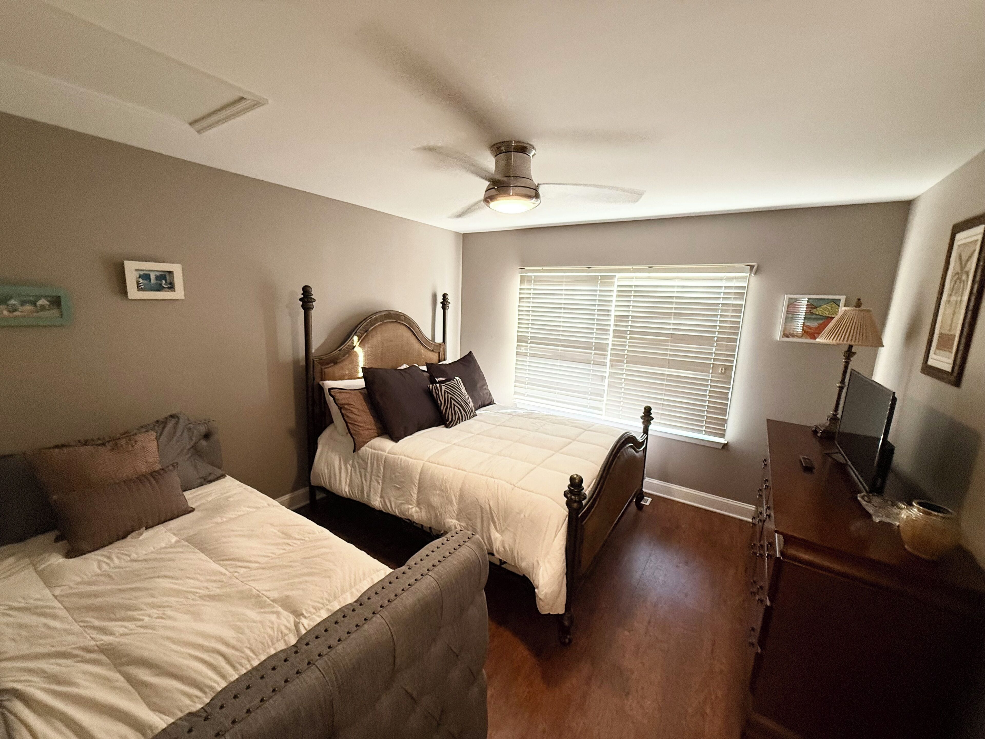 A bedroom with two beds, a ceiling fan, a window with blinds, a bedside table with a lamp, and a framed picture on the wall.
