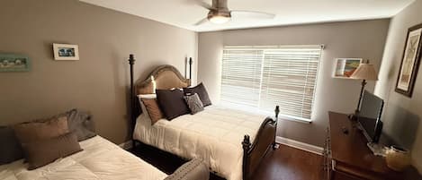 A bedroom with two beds, a ceiling fan, a window with blinds, a bedside table with a lamp, and a framed picture on the wall.