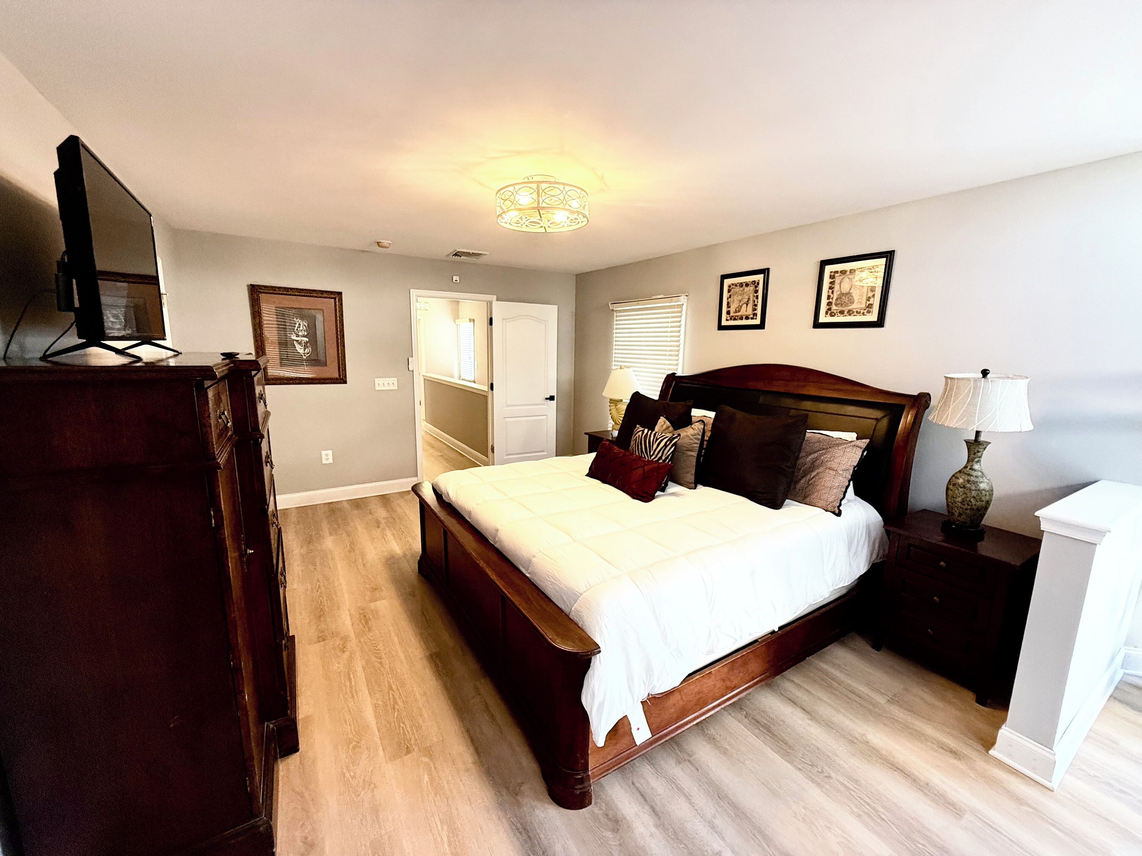 A bedroom with a wooden bed, a dresser, a TV on top, a nightstand with a lamp, and a door leading to another room.