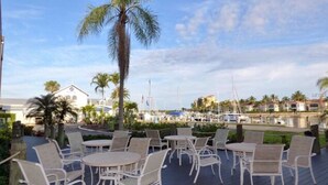 Eine Außenpatio mit weißen Metallstühlen und -tischen, einer Palme und einem Yachthafen mit Booten im Hintergrund.