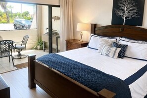 A bedroom with a bed, a nightstand, a lamp, a chair, and a view of the outside.