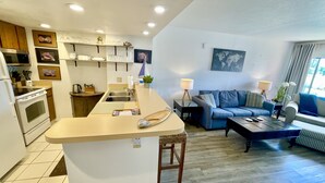 Eine moderne Küche mit Kochinsel, ein Essbereich mit Sofa und Couchtisch sowie ein Wohnzimmer mit Couch und einer Weltkarte an der Wand.