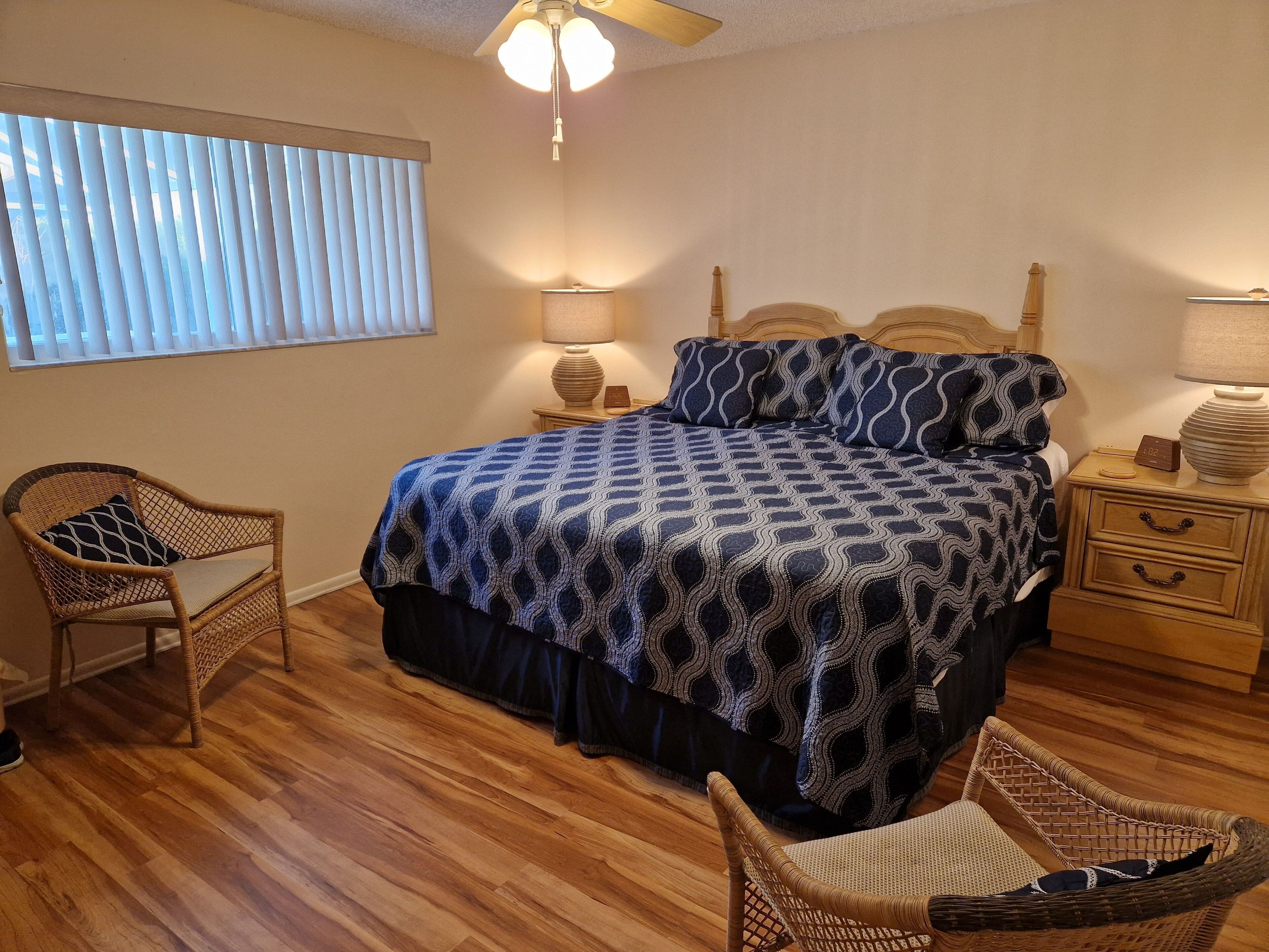 A bedroom with a bed, two nightstands, a ceiling fan, a window with blinds, and two wicker chairs.