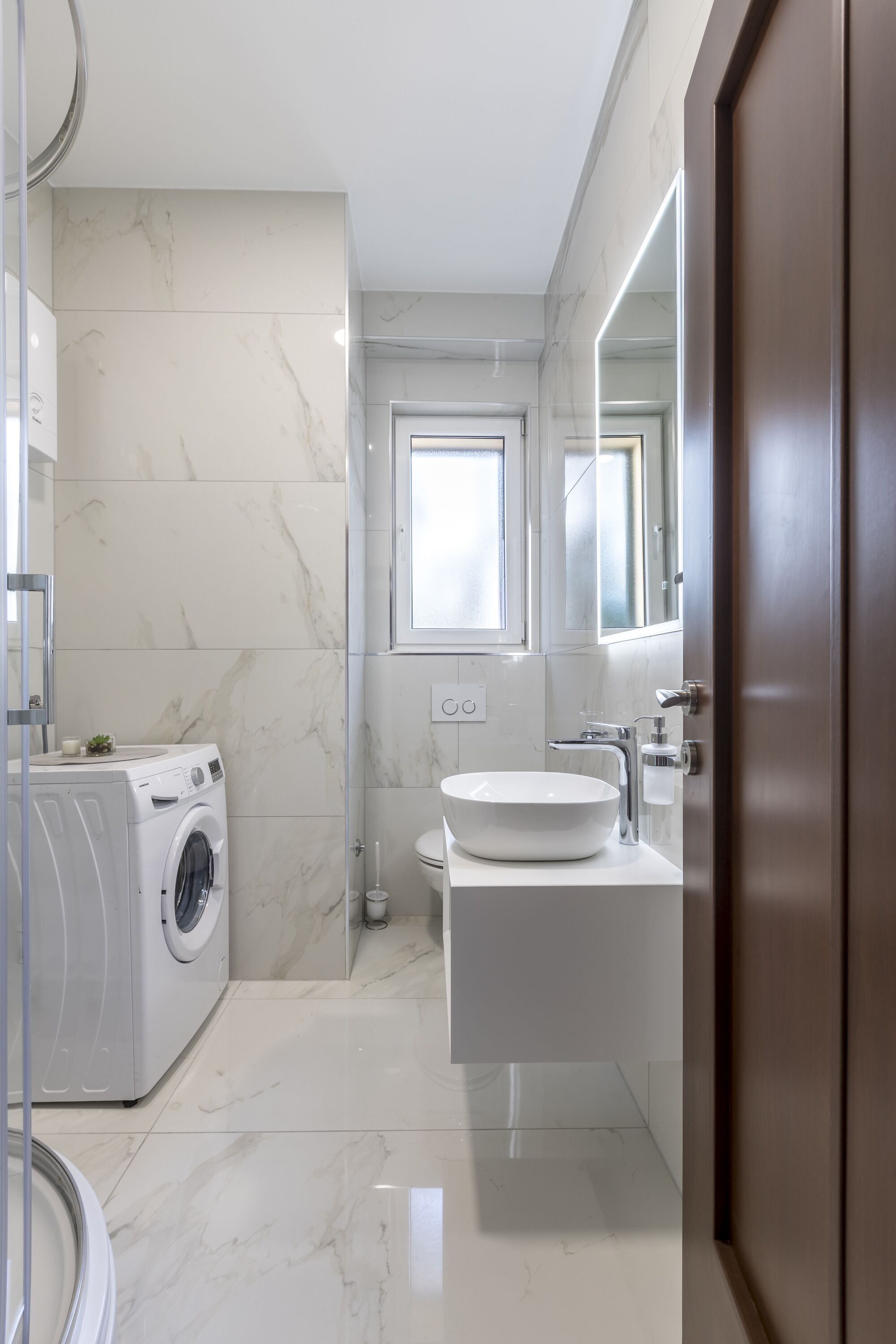 Un baño moderno con piso de mármol, lavadora, lavamanos y un espejo grande.