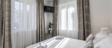 A hotel room with a large bed, white linens, a flat-screen TV mounted on the wall, and sheer curtains.