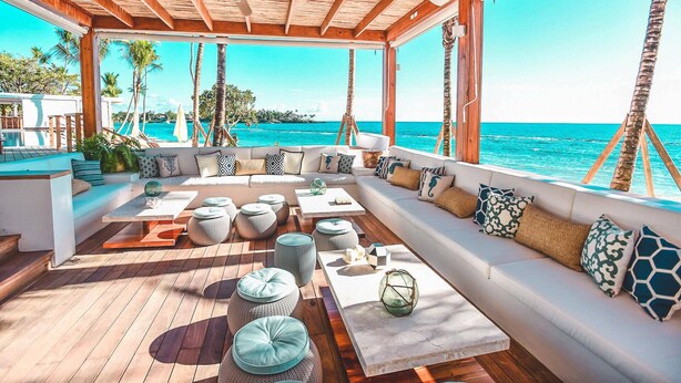 A covered outdoor seating area with a view of the ocean, wooden flooring, and a variety of seating options including sofas and round tables. - Beautiful sea view Golf Villa in Casa de Campo (La Romana)