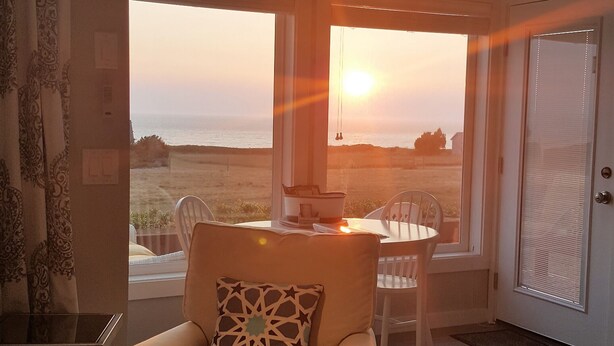 A modern living room with a beige armchair, a dining table with chairs, and large windows offering a view of the sunset over the ocean.