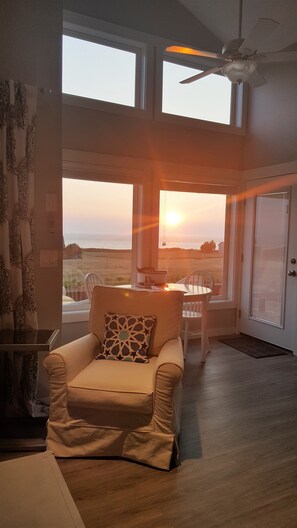 Ein modernes Wohnzimmer mit einem beigen Sessel, einem Esstisch mit Stühlen und großen Fenstern, die einen Blick auf den Sonnenuntergang über dem Meer bieten.
