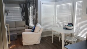 A cozy living area with a sofa, a small table, and a chair. There are patterned curtains and white blinds on the windows.
