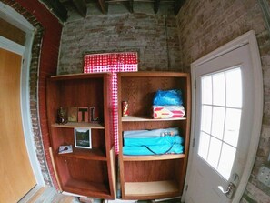 Una habitación con pared de ladrillo, repisa de madera con diversos objetos, ventana con vistas y una puerta.