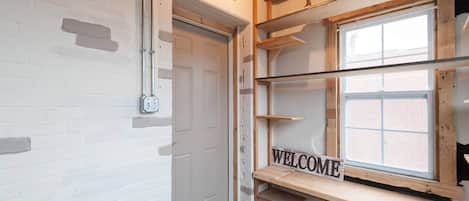 Une pièce avec un mur en briques blanches, une porte fermée, une étagère en bois sur laquelle est posé un panneau indiquant « BIENVENUE », et une fenêtre donnant sur des immeubles.