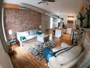 A living room with a sofa, ottoman, and patterned rug. There is a kitchen area with cabinets and a refrigerator. - We do Italian STL style. Perfecto! Enjoy our NBHD! (St. Louis)