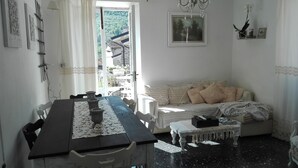A cozy room with a dining table, chairs, a sofa, and a small bench. There is a window with a view of a balcony and trees. - The Haven of Angels (MASSA)