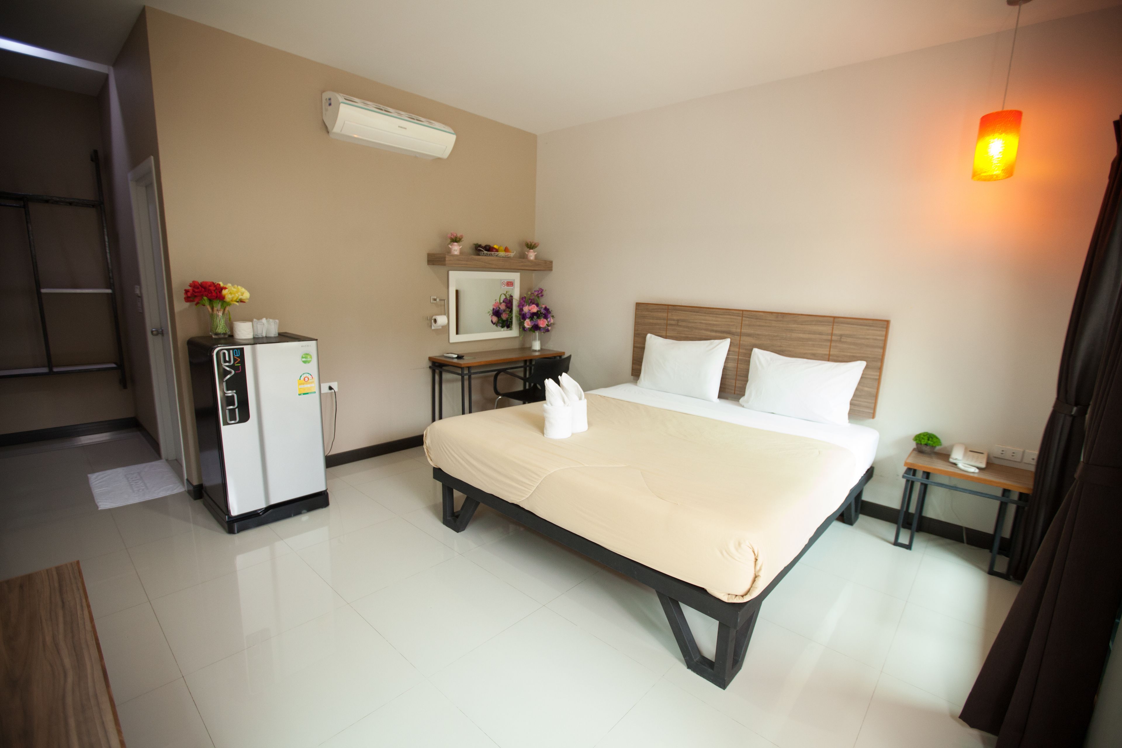 A hotel room with a bed, a desk, a refrigerator, a mirror, and a hanging light fixture.