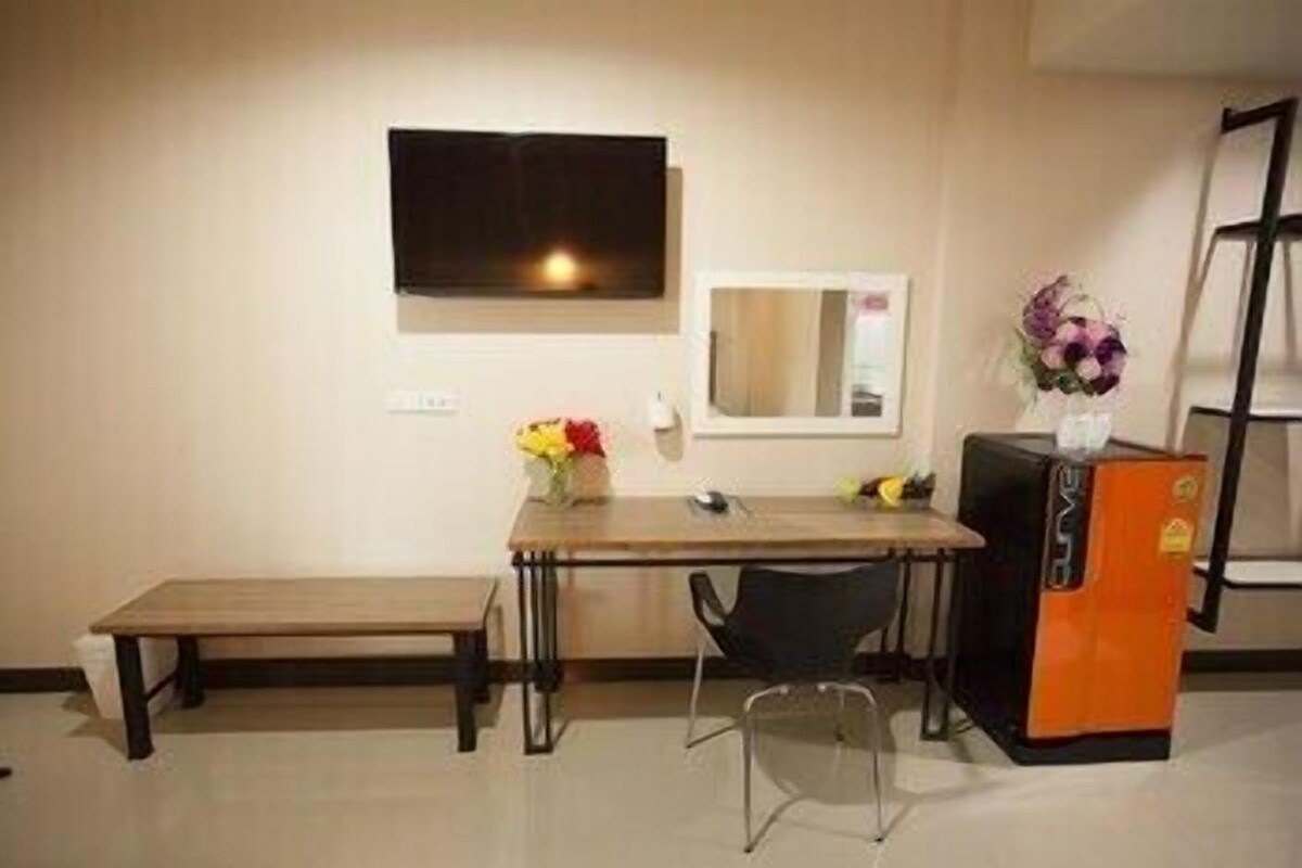 Una habitación pequeña con un escritorio de madera, una silla, un televisor montado en la pared, un espejo y un refrigerador.