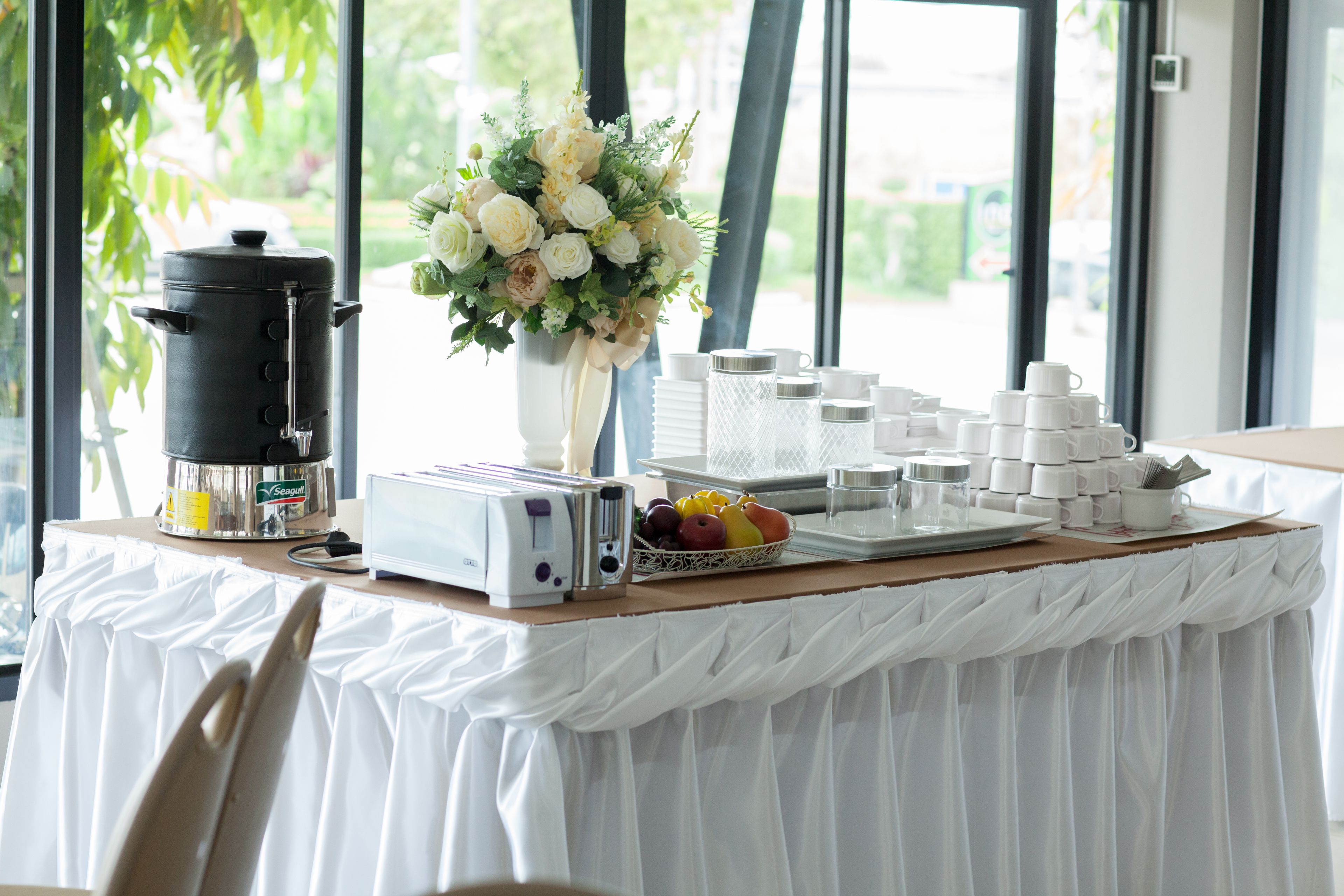 A breakfast setup with a coffee maker, toaster, and fruit bowl on a table with a white tablecloth.