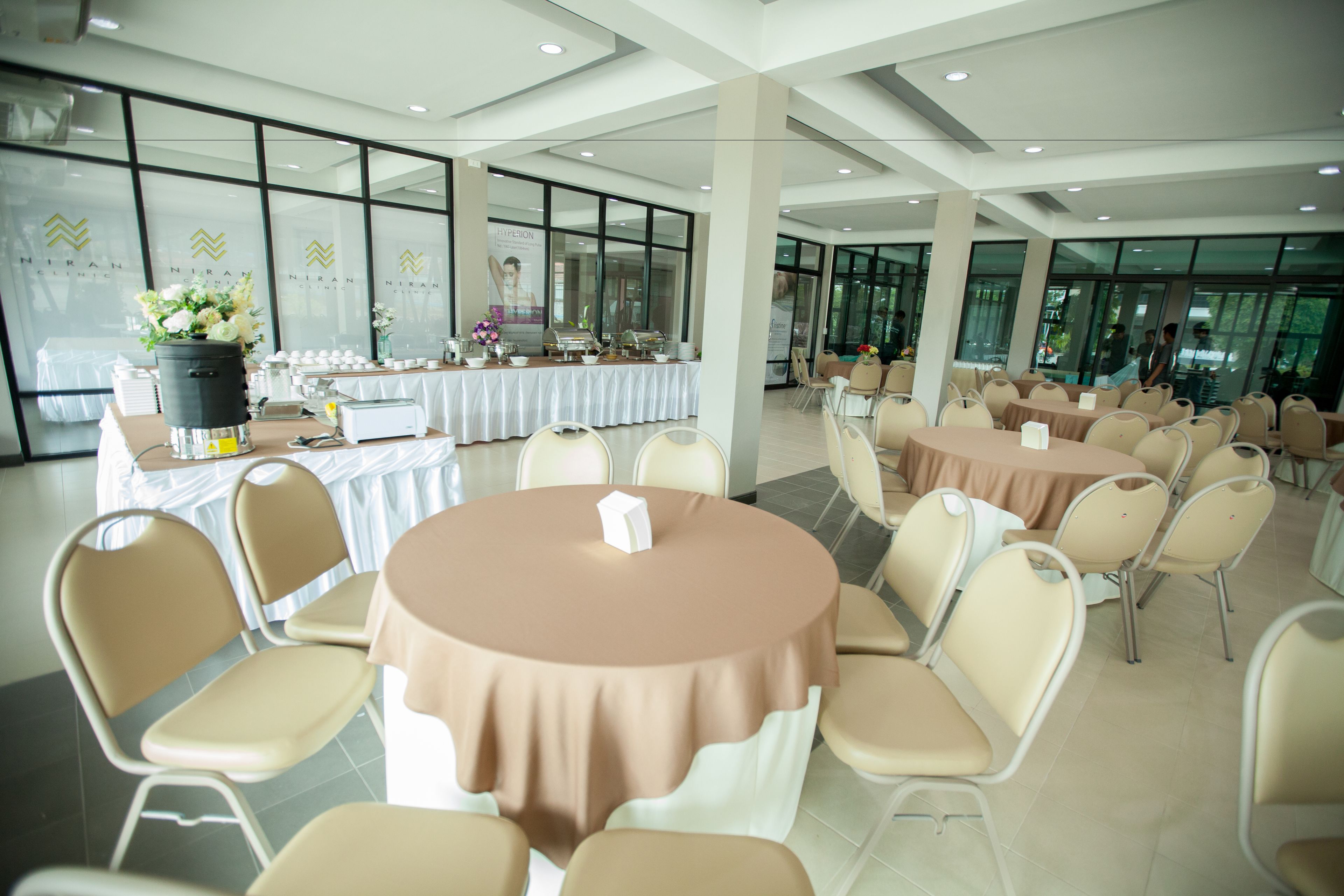 A spacious indoor event space with multiple round tables and beige chairs, large windows, and a buffet setup.