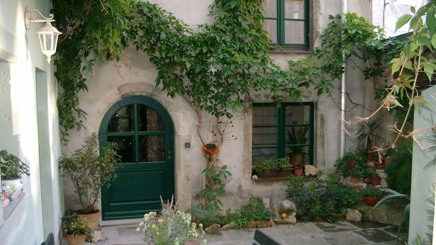 Un patio con una mesa y bancos, rodeado de plantas en macetas y un edificio con puertas y ventanas verdes.