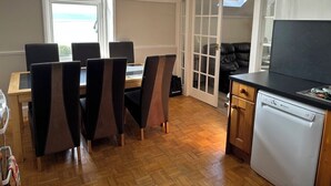 A dining area with a wooden table and chairs, a kitchen with a dishwasher, and a sliding glass door leading to a balcony. - Sealladh na Mara (Gaelic for "View of the Sea") (Isle of Arran)