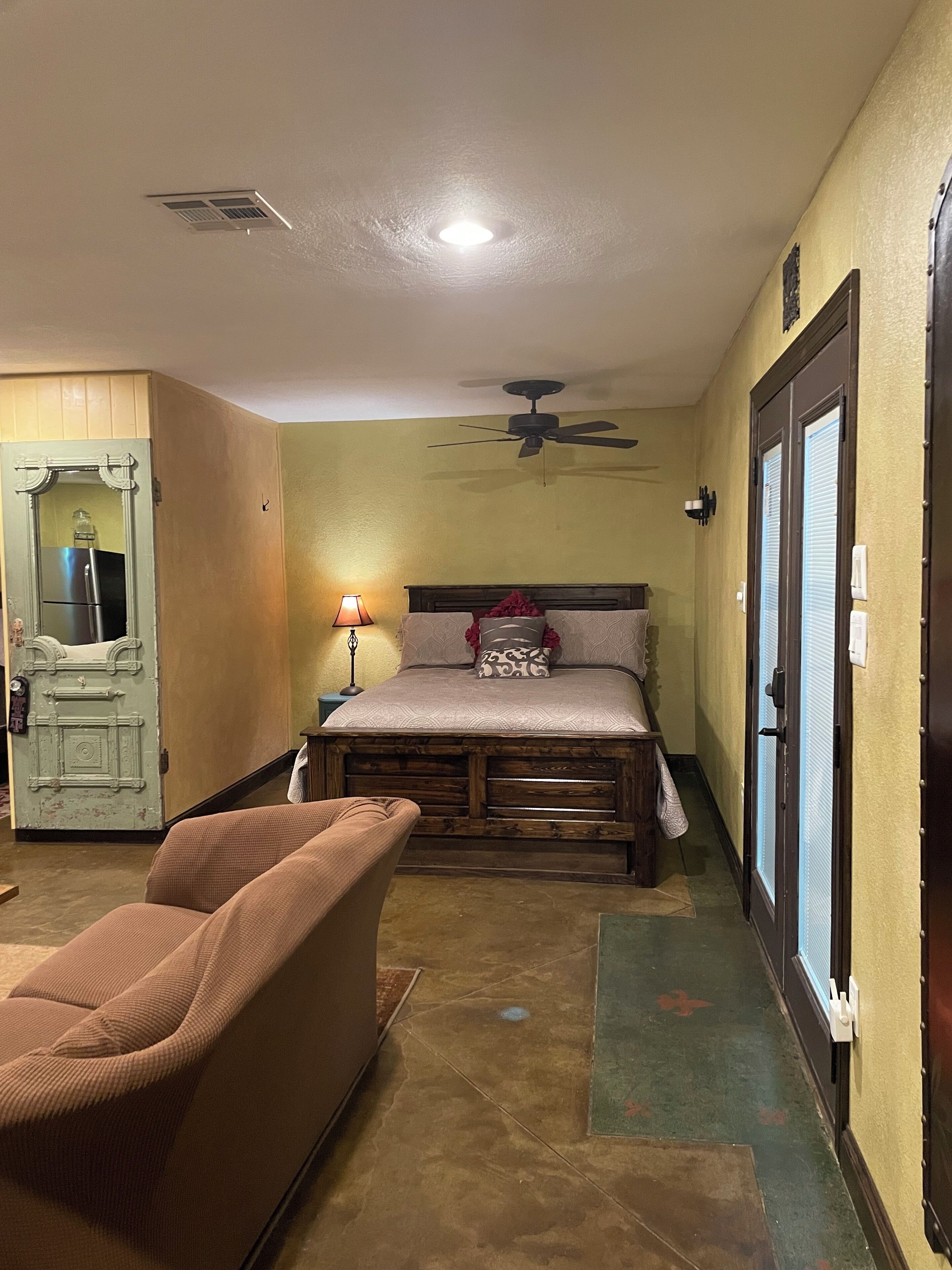 A bedroom with a bed, a ceiling fan, a nightstand, a lamp, and a wooden door.