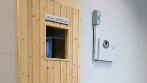 A sauna room with a wooden door, a small window, and a wall-mounted control panel.