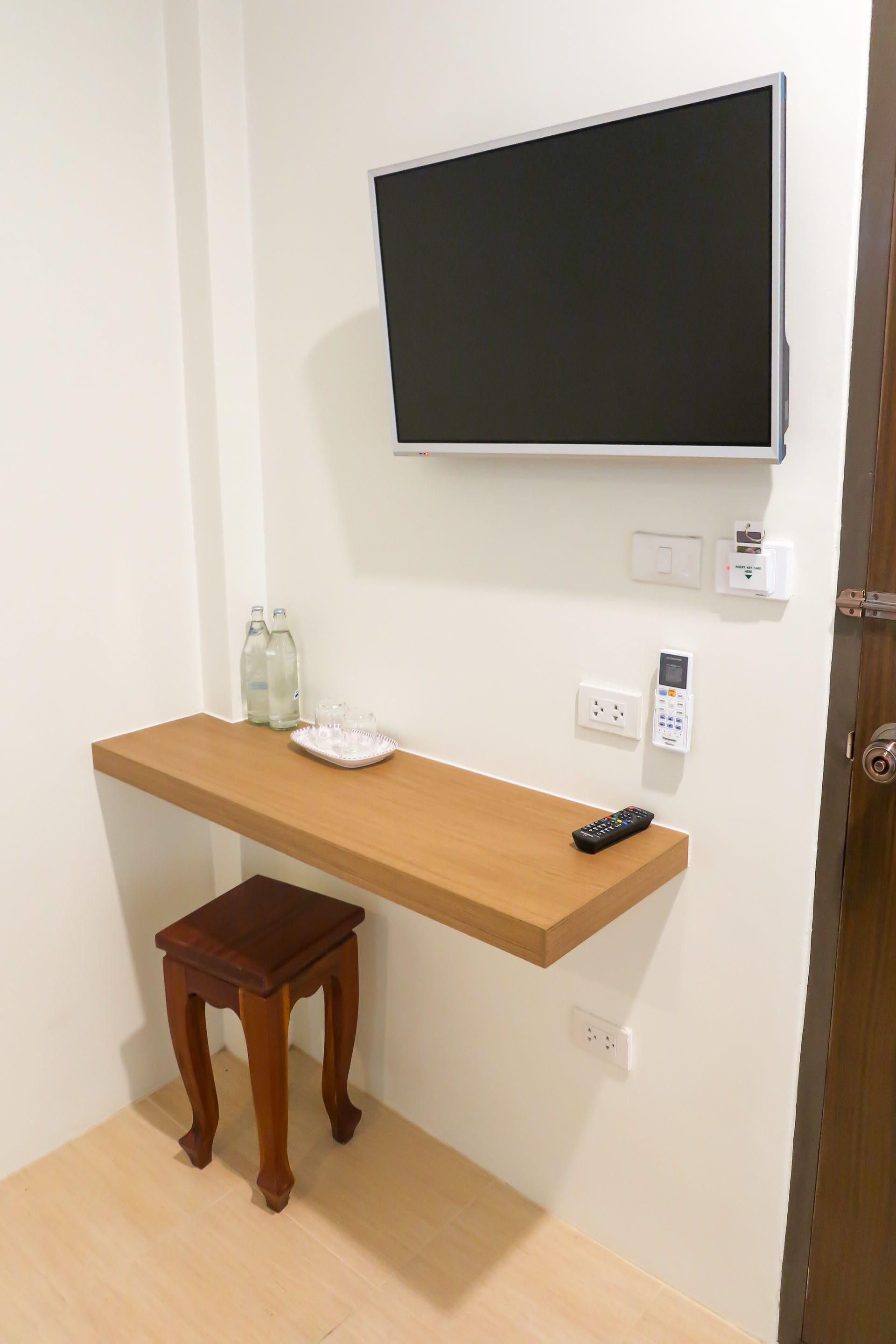 A room with a wooden desk, a television mounted on the wall, a small wooden stool, and a remote control on the desk.