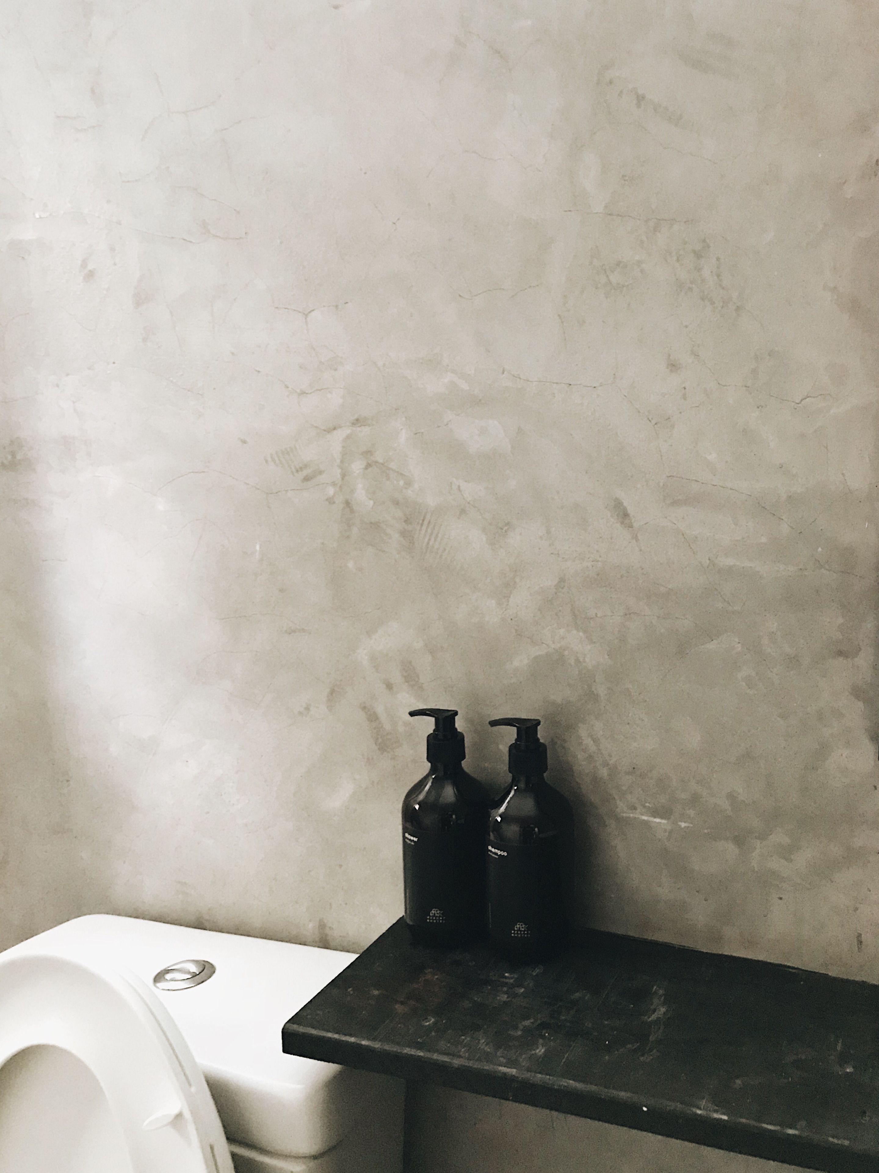 A bathroom with a white toilet, a black shelf with two black bottles, and a textured wall.