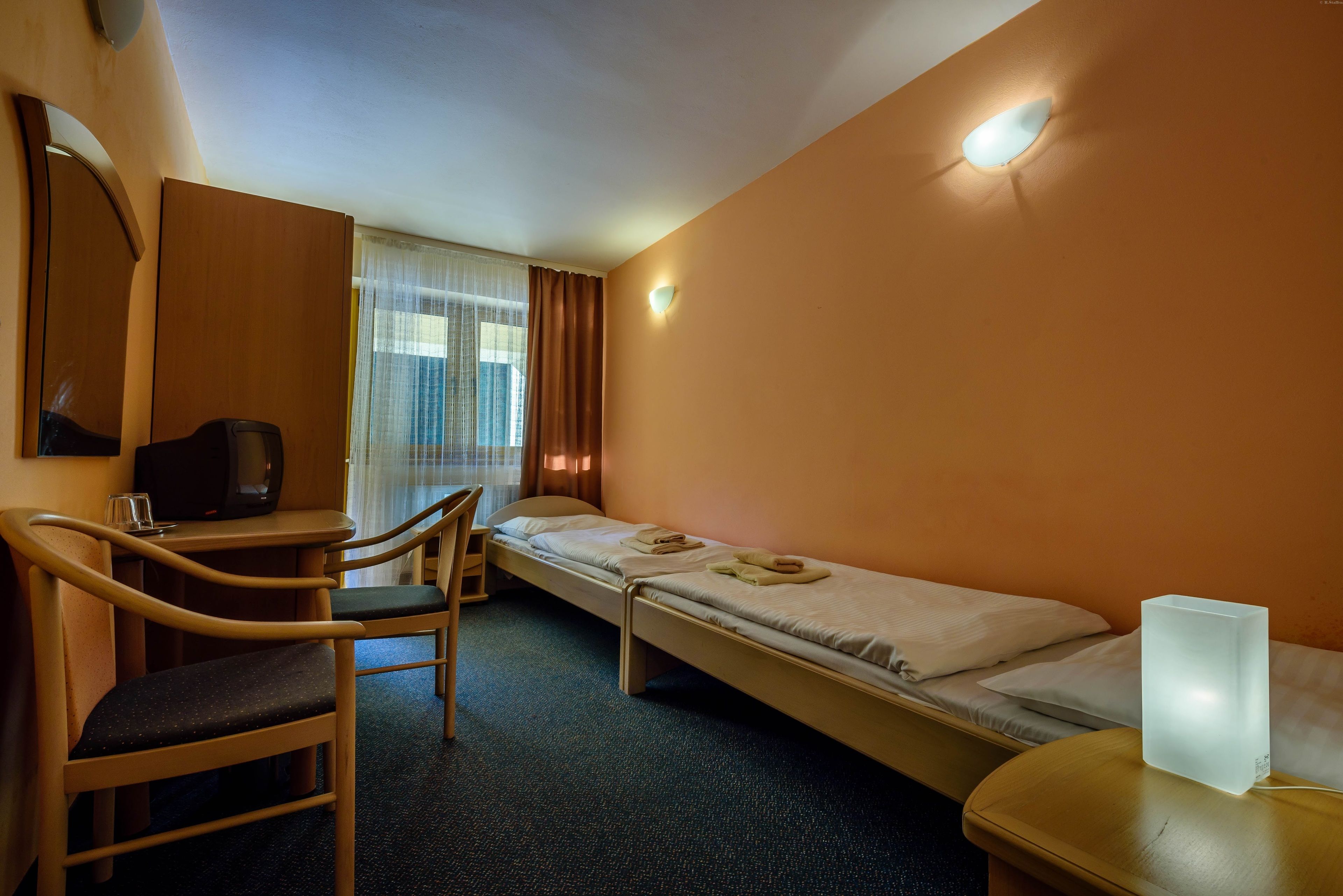 A hotel room with a desk, chair, two single beds, a bedside table, a wall-mounted TV, and a window with curtains.