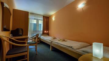 A hotel room with a desk, chair, two single beds, a bedside table, a wall-mounted TV, and a window with curtains.