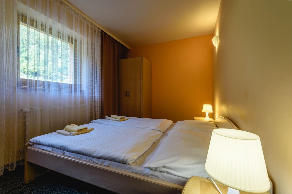 Quarto de hotel com cama, mesa de cabeceira com abajur, guarda-roupa e janela com cortinas translúcidas.