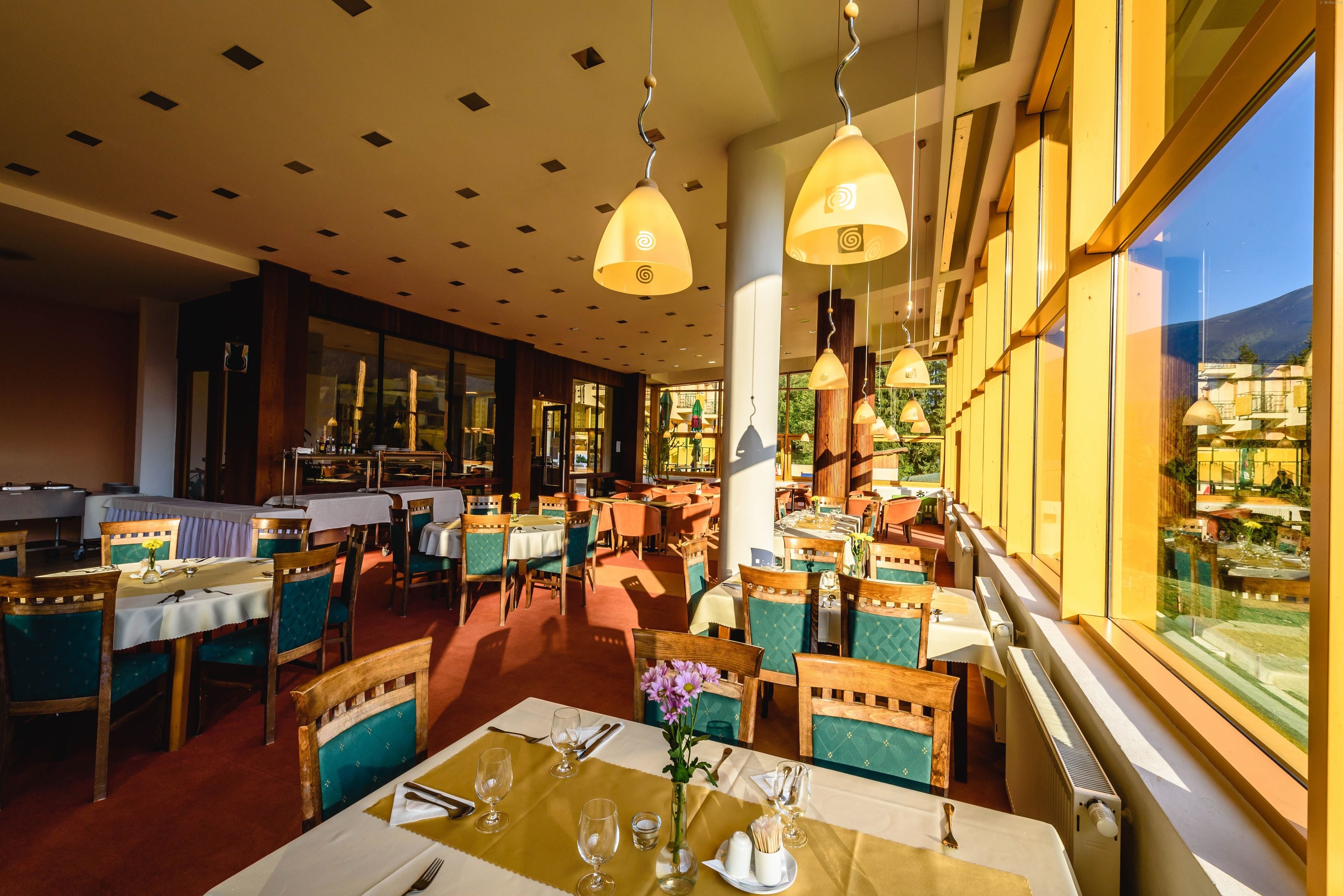 Um restaurante com várias mesas e cadeiras, janelas amplas e luminárias pendentes.