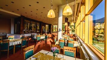 Um restaurante com várias mesas e cadeiras, janelas amplas e luminárias pendentes.