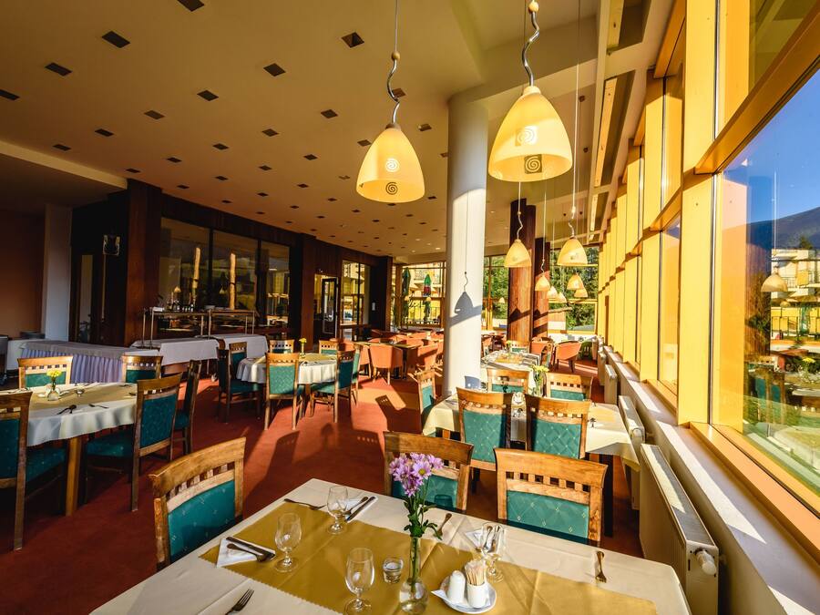 Um restaurante com várias mesas e cadeiras, janelas amplas e luminárias pendentes.