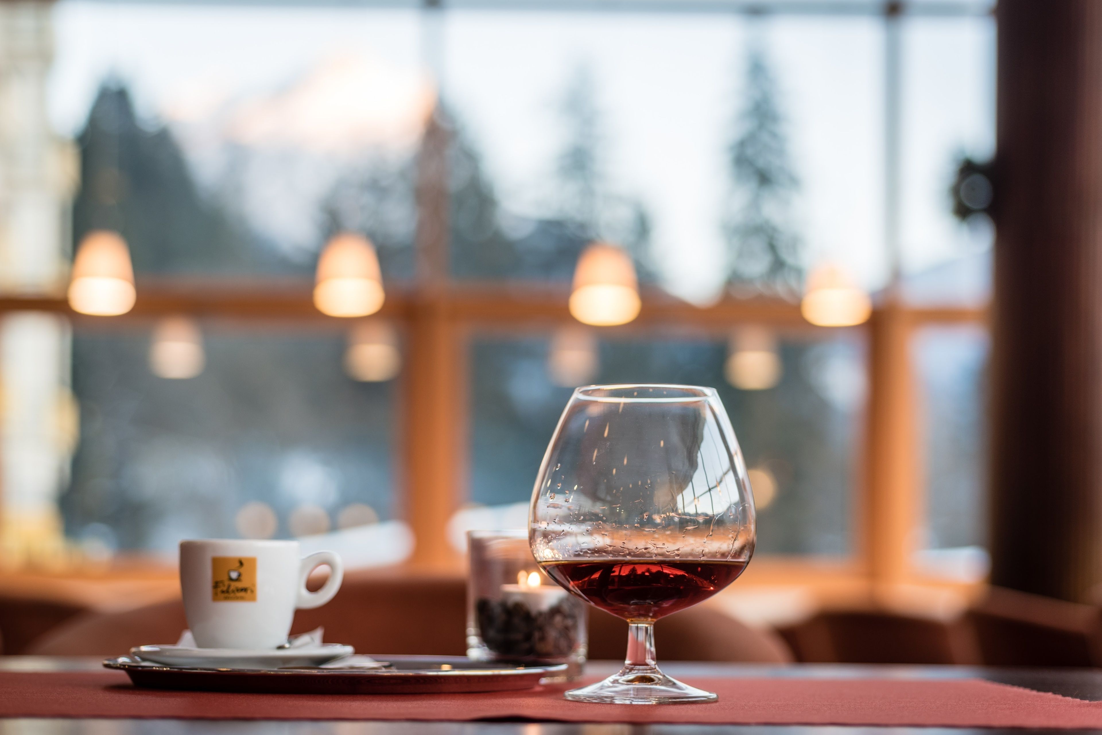 Una mesa con una copa de vino y una taza, una vela, y una vista de un paisaje nevado a través de amplias ventanas.