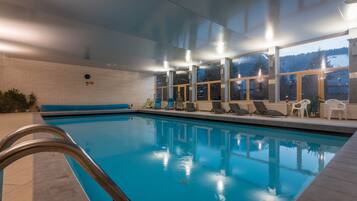 An indoor swimming pool with clear glass walls, a metal pool ladder, and lounge chairs along the side.