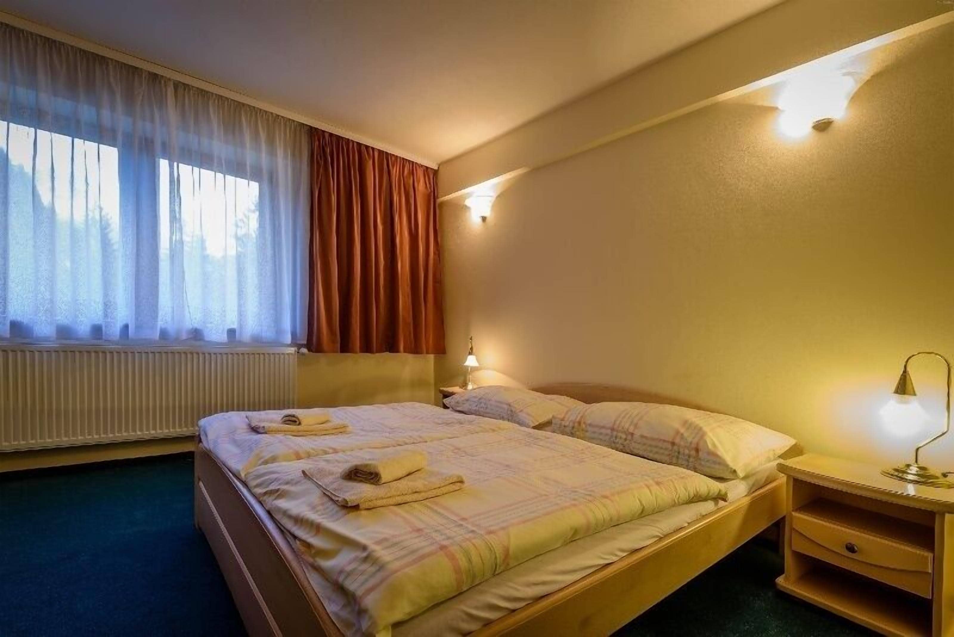 A hotel room with a bed, a nightstand, a lamp, a window with curtains, and a wall-mounted light.