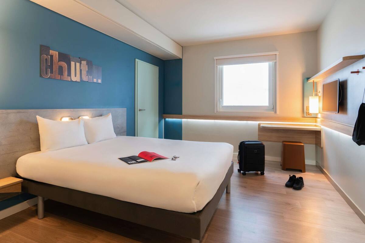 Habitación de hotel con cama, mesita de noche, escritorio, silla, maleta y televisor.