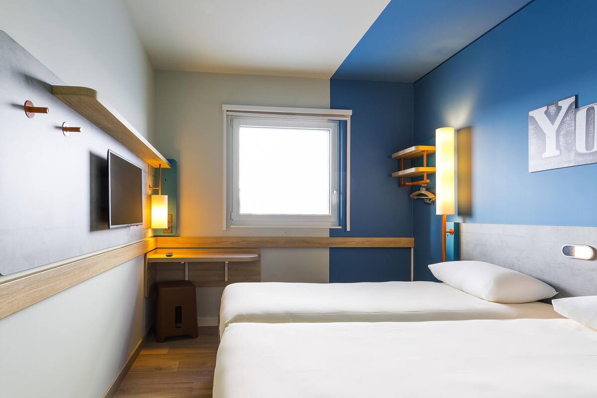 Habitación de hotel con dos camas, un escritorio, un televisor, una ventana y un estante con luces.