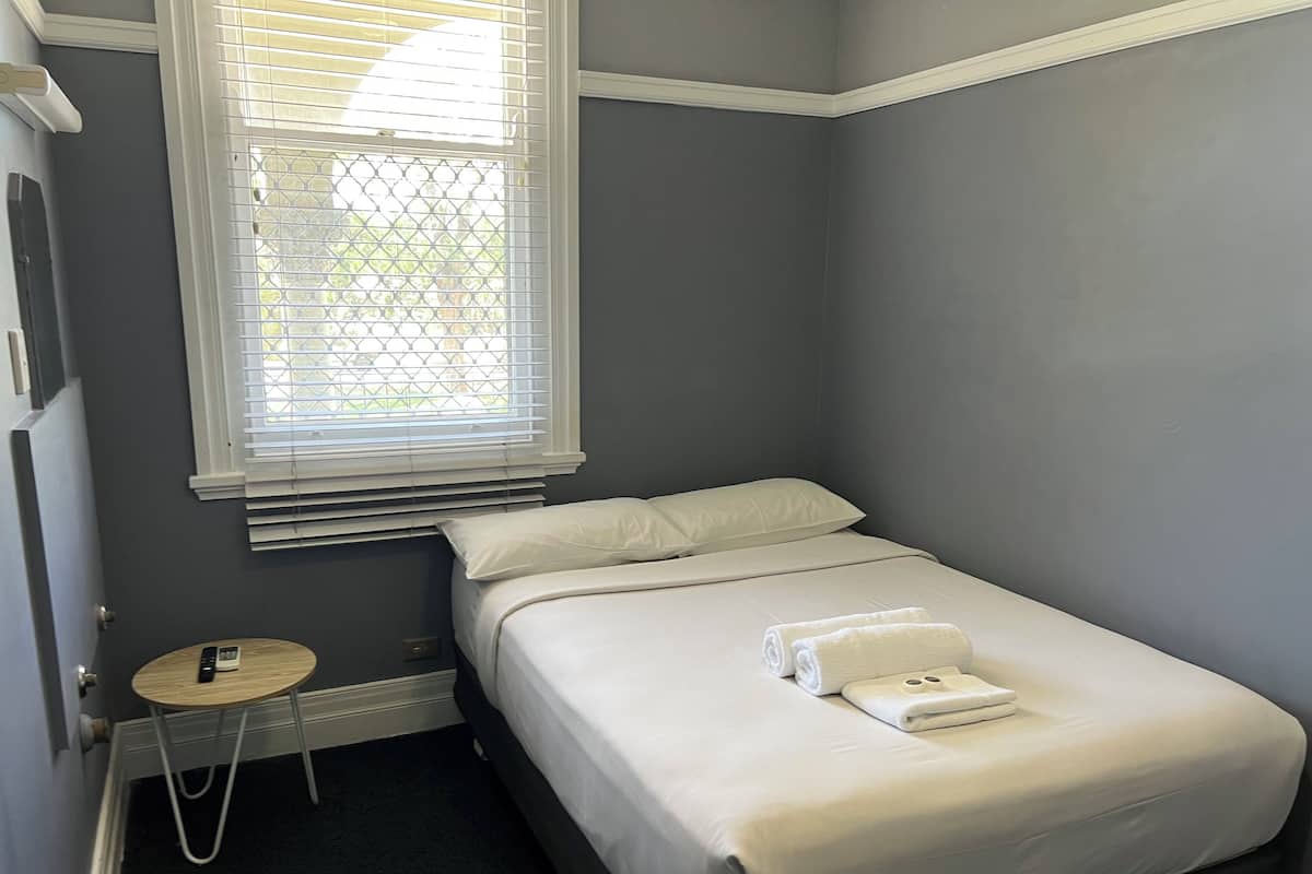 A small, neatly arranged bedroom with a single bed, a bedside table with a remote control, and a window with blinds.