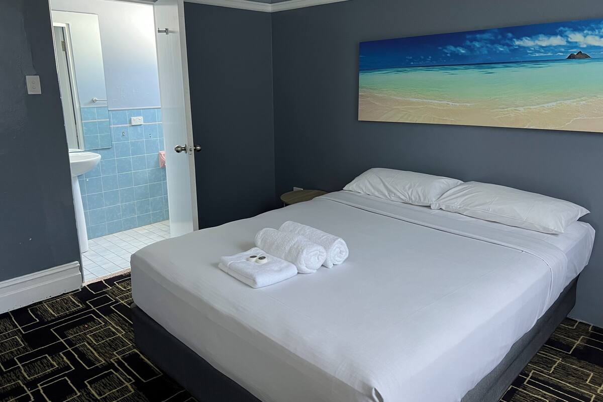 A hotel room with a bed, a painting of a beach, a bathroom with blue tiles, and a patterned floor.