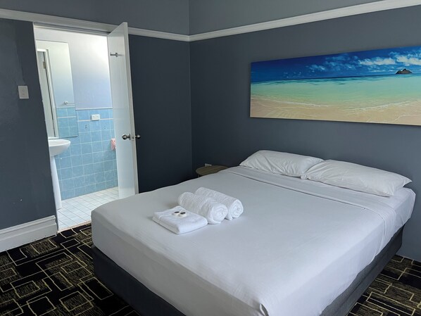 A hotel room with a bed, a painting of a beach, a bathroom with blue tiles, and a patterned floor.