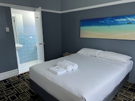A hotel room with a bed, a painting of a beach, a bathroom with blue tiles, and a patterned floor.