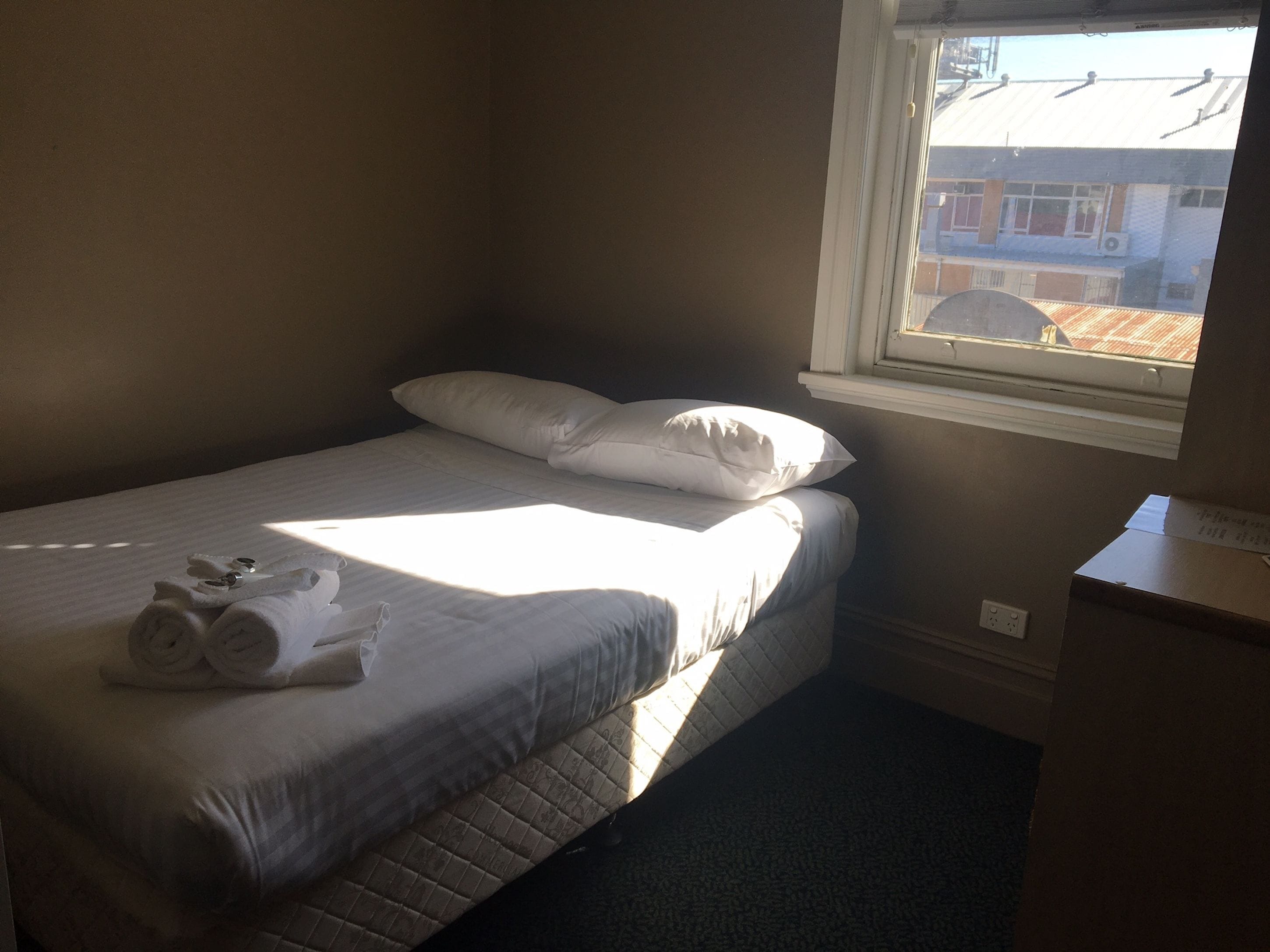 A single bed with white linens, a window with blinds, and a small desk with a chair.