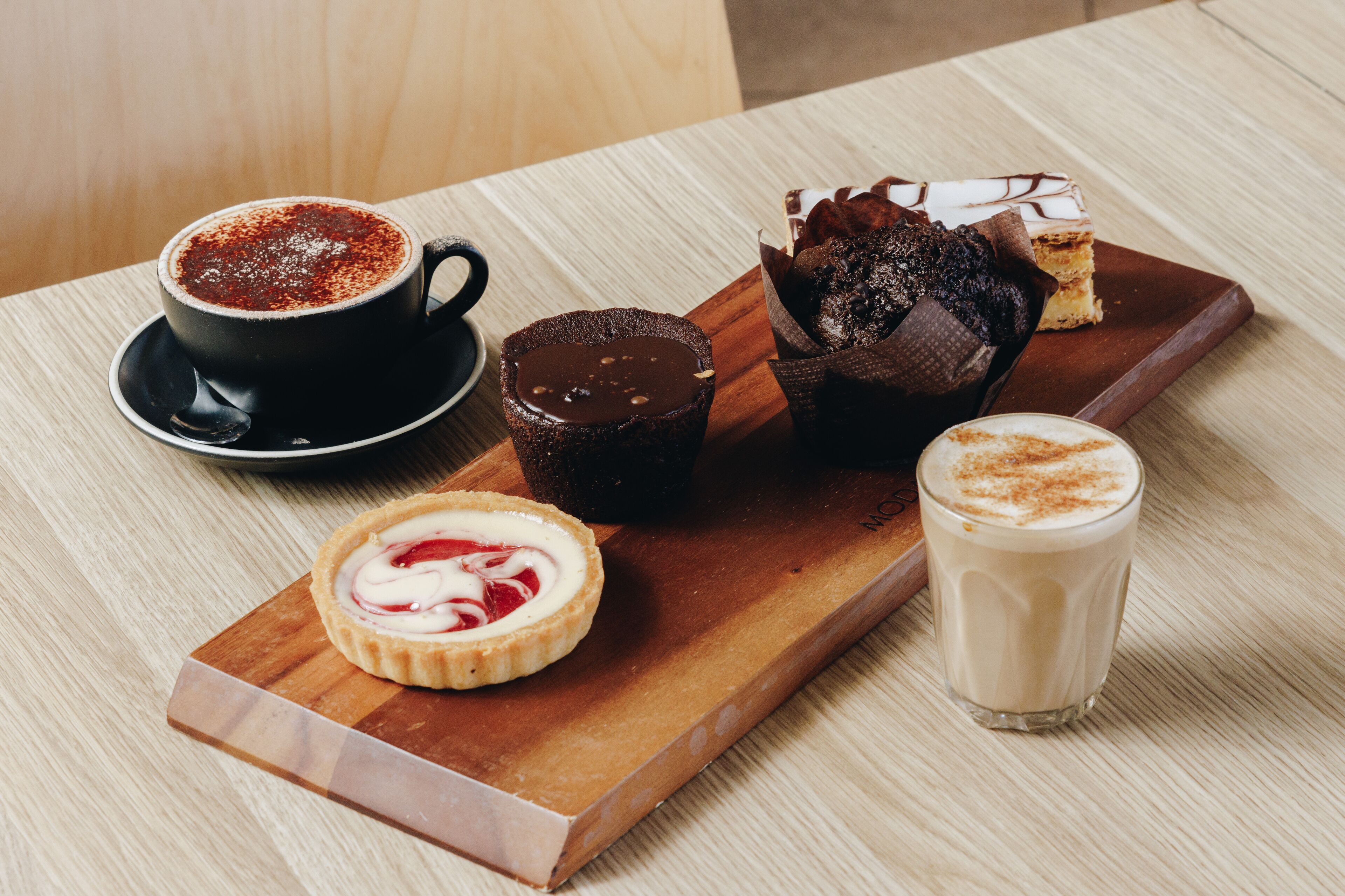 A wooden board with a cup of coffee, a chocolate dessert, a muffin, and a tart on a wooden table.