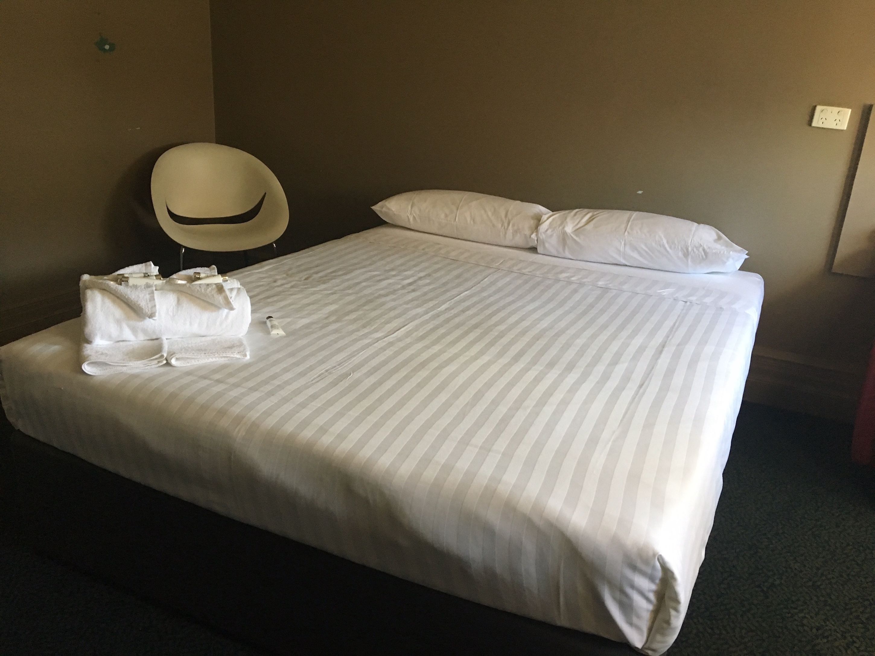 A hotel room with a neatly made bed, a round chair, and a white pillow.